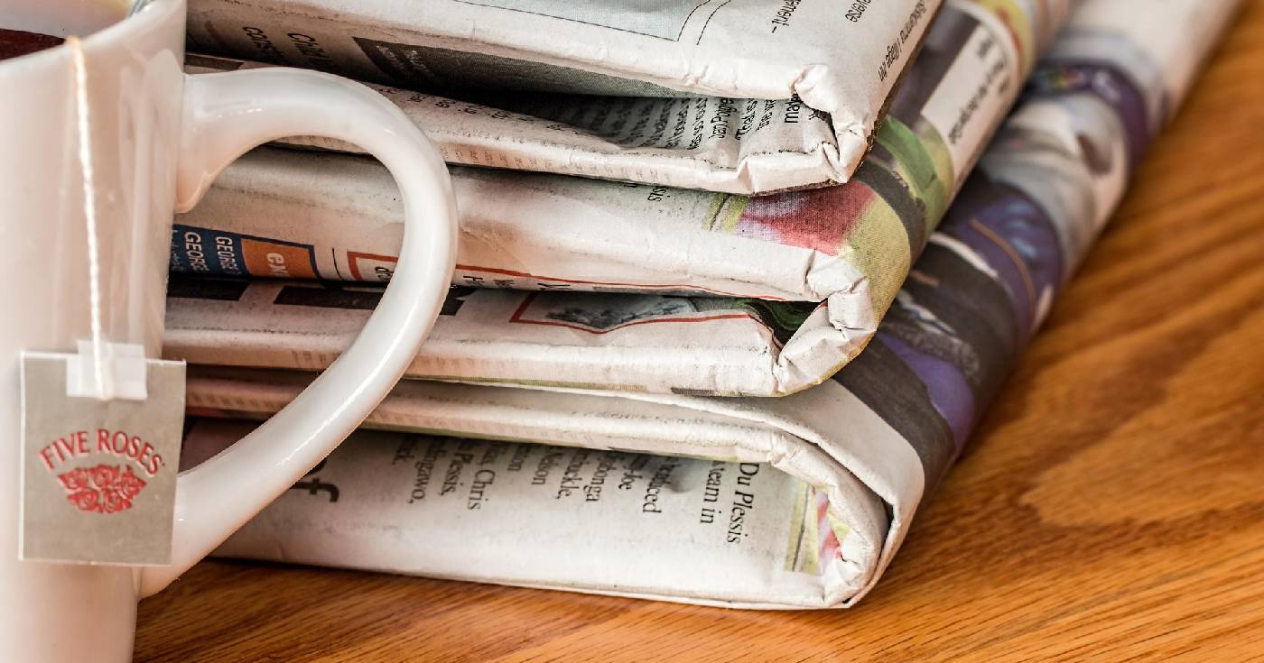 A stack of newspapers sits on a wooden table next to a cup of tea with a teabag hanging out of it.